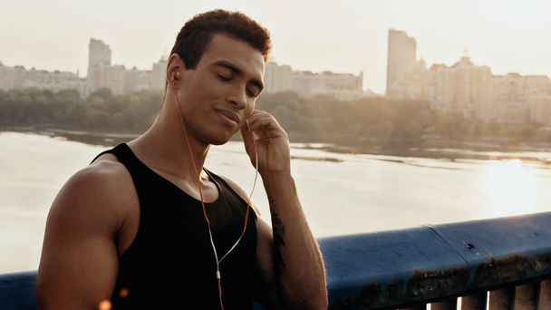 uśmiechnięty mieszany wyścig człowiek z zamkniętymi oczami słuchanie muzyki w słuchawkach na moście nad rzeką - Zdjęcie, obraz