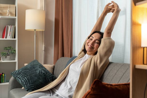 Позитивная молодая азиатка, сидящая дома на диване и закрывая глаза, растягиваясь утром. Фото на складе - Фото, изображение