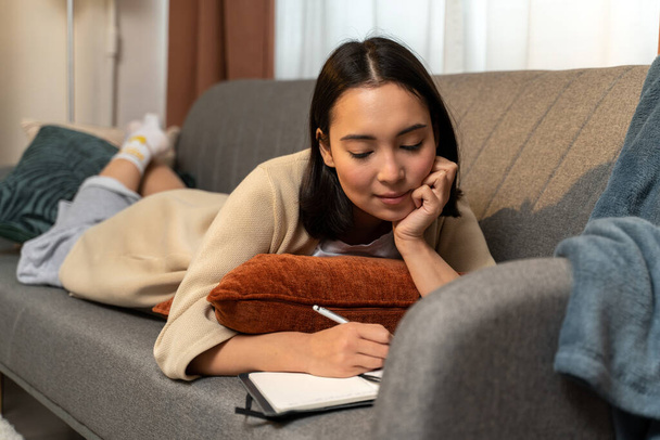 Pełna długość widok azjatyckiej kobiety relaks na kanapie w domu podczas pisania notatek do notatnika. Zdjęcie z magazynu - Zdjęcie, obraz