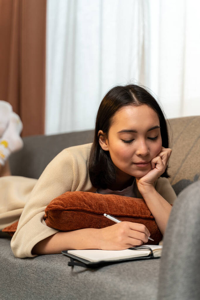 Thoughtful young asian woman dreaming while writing notes at home at the morning alone. She laying at the sofa. Stock photo - Fotó, kép