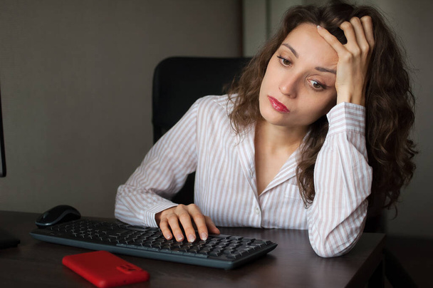 Giovane donna stanca con i capelli ricci e camicia bianca sta lavorando in ufficio utilizzando il suo computer portatile, lavoro di routine, freelance, sindrome di burnout - Foto, immagini