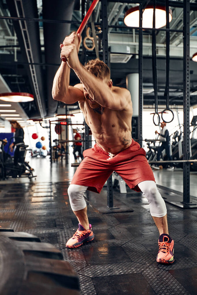 Strength concept. Male athlete using hammer during fitness workout. Fitness man beating rubber tire in gym. Muscular sportsman performing intensity training with sledgehammer in sport club - Photo, Image