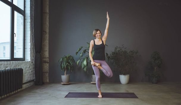 Lonely Woman in Sportswear Doing Side Plank in a Spacious Yoga Studio. Sporty Fit Woman Practices Hatha Yoga. Full Length. Gray Background - 写真・画像