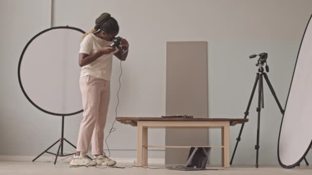 Apuñalada de cuerpo entero de una joven fotógrafa afroamericana tomando fotos en una cámara digital en un estudio fotográfico con iluminación y equipo profesional - Metraje, vídeo
