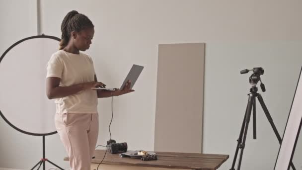 Μεσαία φωτογραφία Αφρο-Αμερικανίδας φωτογράφου φαγητού που δουλεύει σε laptop και φωτογραφίζει όμορφο πρωινό σε ψηφιακή κάμερα, δημιουργώντας περιεχόμενο σε φωτεινό φωτογραφικό στούντιο - Πλάνα, βίντεο