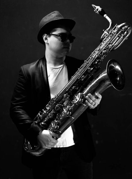Black and White. Young Saxophonist in a Black Jacket, Hat and Sunglasses Holds a Baritone Saxophone. Dark Background. Neon Light. Close-up Portrait - Foto, Bild