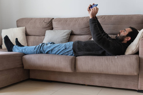 Brown caucasian man with a beard, lying on a sofa while interacting with his blue smart phone - Foto, afbeelding