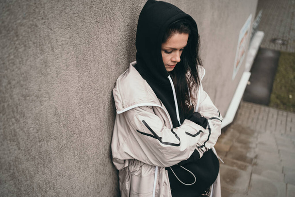 An upset girl stands against a wall on the street. A girl in a black hoodie. Space for text. A teenager in a black hood is offended.  - Valokuva, kuva