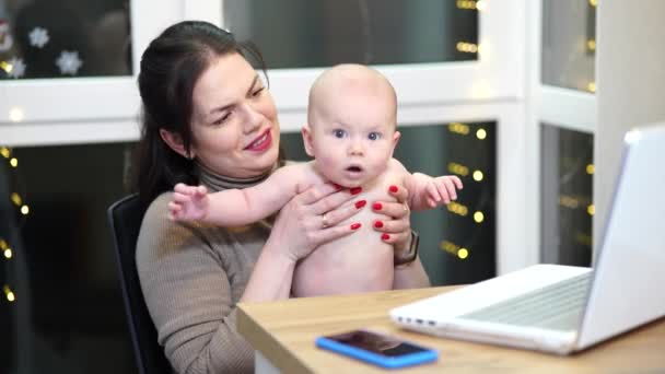 Mujer joven con niño pequeño en brazos escribiendo en el ordenador portátil, trabajo remoto con un bebé en casa. - Metraje, vídeo