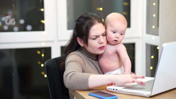 Junge Frau mit Kleinkind im Arm tippt auf Laptop, Fernarbeit mit Baby zu Hause. - Filmmaterial, Video