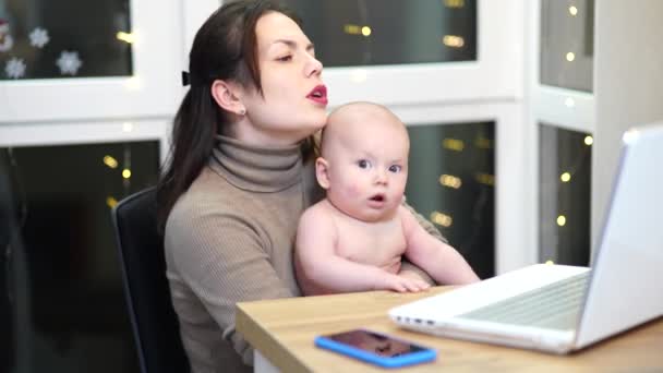Young woman with toddler in arms typing on laptop, remote work with a baby at home. - Filmagem, Vídeo