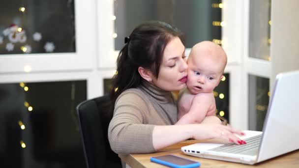 Молода жінка з малюком на руках друкує на ноутбуці, віддалена робота з дитиною вдома
. - Кадри, відео