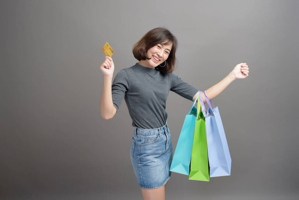 A portrait of young beautiful asian woman holding credit card and colorful shopping bag isolated over gray background studio - Photo, Image