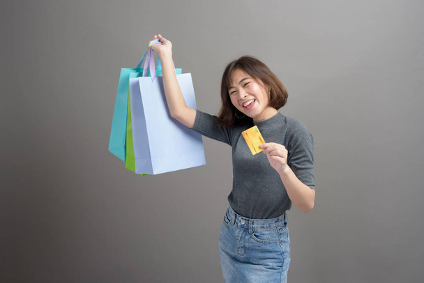 A portrait of young beautiful asian woman holding credit card and colorful shopping bag isolated over gray background studio - Fotoğraf, Görsel