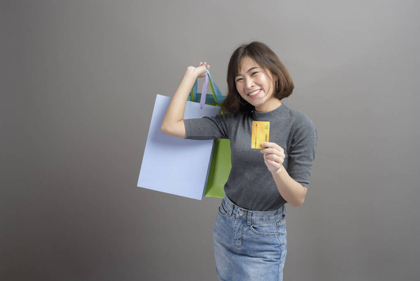 A portrait of young beautiful asian woman holding credit card and colorful shopping bag isolated over gray background studio - Fotó, kép