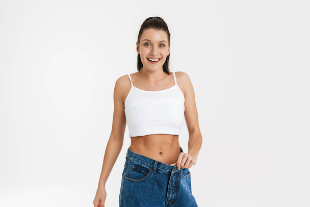 European brunette woman smiling while posing in big jeans isolated over white background - Zdjęcie, obraz