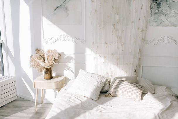 Стильный и уютный интерьер спальни. Ваза с сушеными цветами на тумбочке в светлой спальне, солнце светит из окна на кровати - Фото, изображение