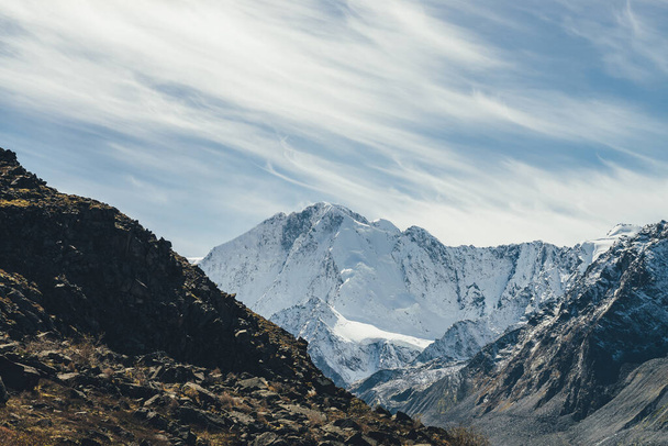 Alpejski krajobraz z wysoką zaśnieżoną górą ze szczytowym szczytem pod chmurami cyrru na niebie. Atmosferyczny widok na duże góry pokryte śniegiem w słońcu. Czarne skały i biały śnieg spiczasty szczyt w świetle słonecznym. - Zdjęcie, obraz