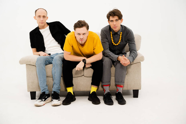 Young Guys Sit on a Sofa on a White Background and Look Together at the Camera. Three Friends are Sitting on the Couch and Watching TV, Watching a Home Theater Together. Close-up - Foto, afbeelding