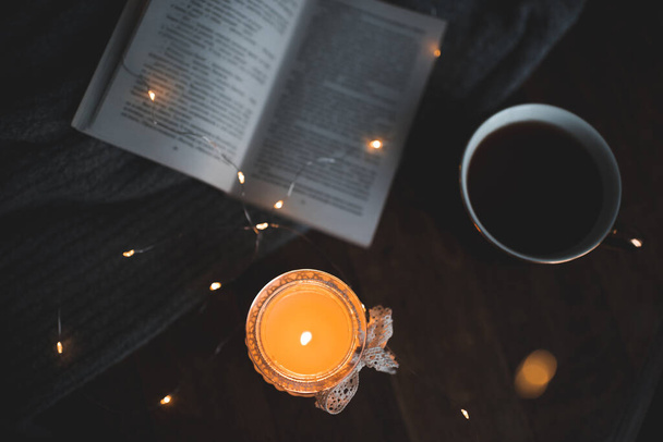 Dark and cozy atmosphere in room: open paper book with burning candle and cup of fresh coffee on wooden table closeup top view at home. Reading at night.  - Photo, Image