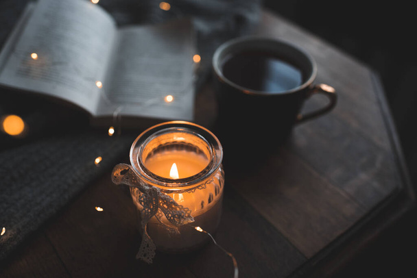 Tmavá a útulná atmosféra v pokoji: otevřená papírová kniha s hořící svíčkou a šálek čerstvé kávy na dřevěném stole detailní up doma. Čtení v noci.  - Fotografie, Obrázek