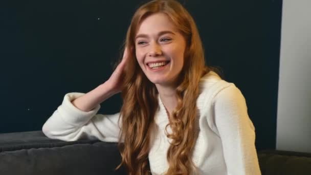 Adorable woman with ginger hair and freckles is sitting on the couch and smile at camera - Séquence, vidéo