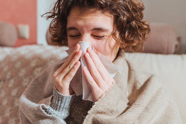 Хвора засмучена молода жінка сидить на дивані вкритому теплим пледом, замерзає, похмуро, нежить, гарячка, чхає в тканину, хвора дівчина з симптомами грипу, кашляє вдома, концепція грипу
 - Фото, зображення