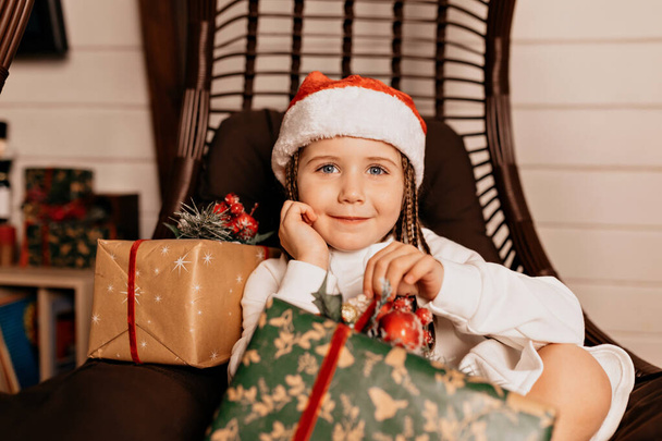 Adorable happy kid in Santa cap holding her big present and truly smiling at camera. Holiday mood, birthday party. New Year, Christmas . High quality photo - Photo, Image