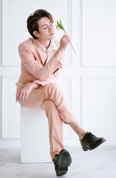 Smart young casual Asian man sitting on white cubes and holding white tulip. Business portrait of young Korean man in pink suit and looking flower at studio background. - Фото, зображення