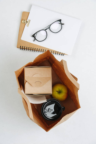 Top view composition of a lunch in a paper bag and glasses on a notepad. Apple, cup of coffee and a sealed paper box with a meal. Eco friendly packaging concept. Over white background. - Фото, изображение