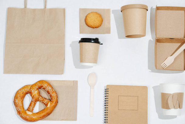 Composition of eco friendly items arranged in a flat lay. Carton compostable products over white. Cups, box, bag, cutlery, notepad completed with some pastry. - Φωτογραφία, εικόνα