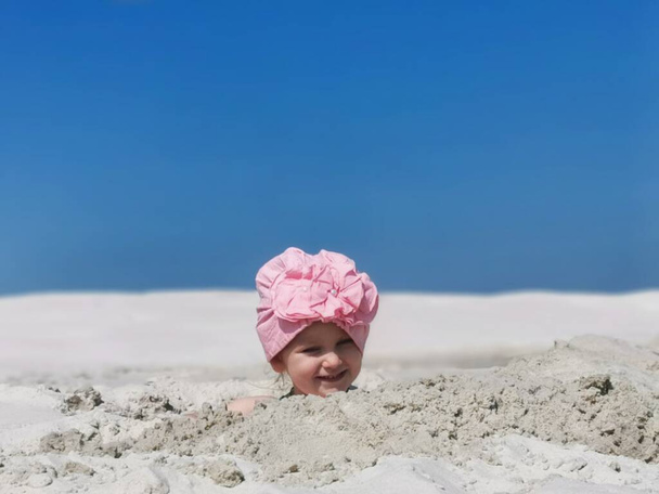 malé dítě si hraje v písku na pláži, pohřbeni až k hlavě, dívka v šále se usmívá při hraní, bílý písek, léto, modrá obloha, horizontální fotografie, rodinná dovolená, hry s dětmi - Fotografie, Obrázek