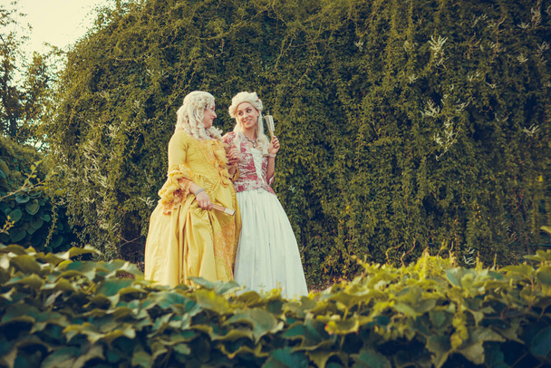 Portret dwóch blond kobiet ubranych w historyczne stroje barokowe ze starą fryzurą mody, na świeżym powietrzu. Luksusowa średniowieczna sukienka - Zdjęcie, obraz