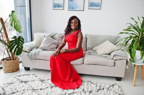 Magnificent young african woman in luxurious red dress in a luxury apartment sitting at couch. Beauty, fashion. - Photo, Image