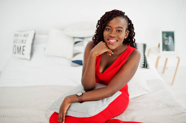 豪華なアパートのベッドの上に座って豪華な赤いドレスで壮大な若いアフリカの女性。美、ファッション. - 写真・画像
