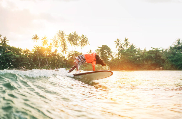 Black long-haired teen boy riding and jumping from a long surfboard. He caught a  wave in an Indian ocean bay with magic sunset background. Extreme water sports and exotic countries concept. - 写真・画像