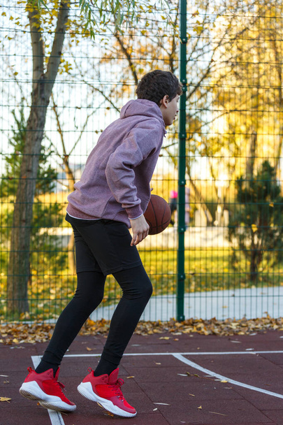 Cute teenager in a hoodie playing basketball. Young boy with ball learning dribble and shooting on the city court. Hobby for kids, active lifestyle - Fotografie, Obrázek