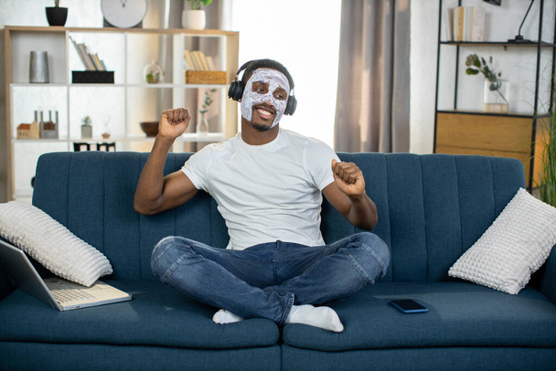 Χαλαρωμένος Αφροαμερικάνος με λευκή μάσκα από πηλό, κάθεται στον καναπέ στο σπίτι και ακούει μουσική - Φωτογραφία, εικόνα