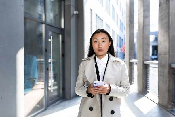 Красива азіатська бізнес-леді дивиться на камеру і посміхається, тримаючи телефон в руках, біля офісного центру зовні
 - Фото, зображення