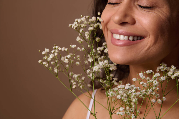 Close-up portrait of Hispanic pretty woman with closed eyes and beautiful smile, holding Gypsophila white sprig against beige background. Femininity, sensuality, natural beauty, Women's Day concept - Photo, Image