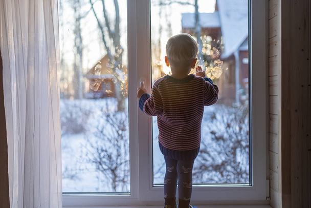 little boy stand near the window and look at the snowy street in winter, when it is very cold. - Foto, immagini