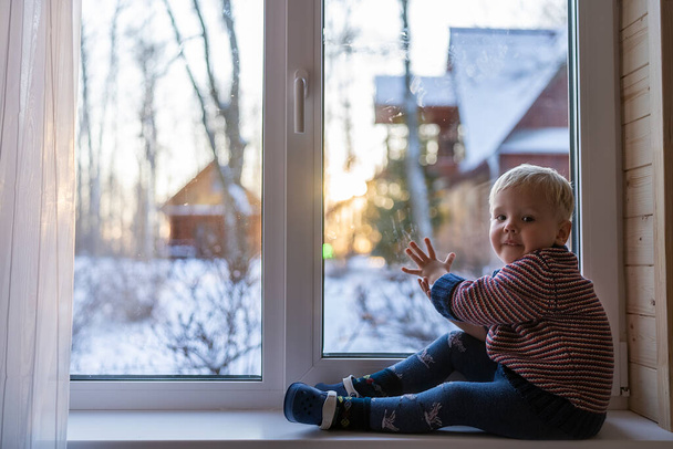 little boy stand near the window and look at the snowy street in winter, when it is very cold - Фото, изображение