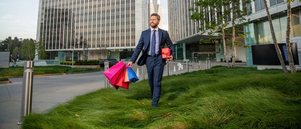 hombre feliz y exitoso en ropa formal con bolsas de compras y caja presente fuera de la oficina, negocio. - Foto, Imagen
