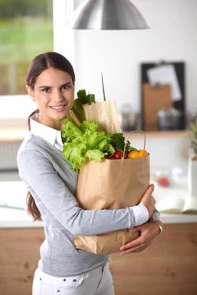 野菜と食料品の買い物袋を持っている若い女性キッチンに立つ. - 写真・画像