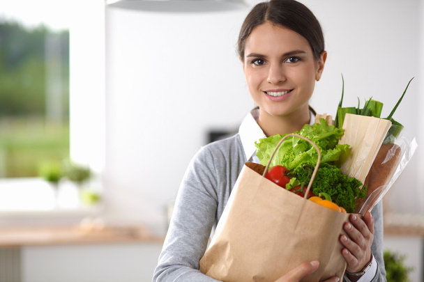 Jovem segurando supermercado saco de compras com legumes de pé na cozinha. - Foto, Imagem