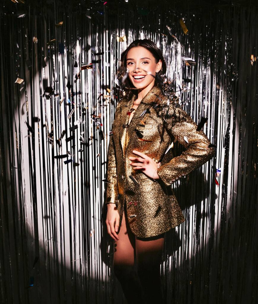 Young beautiful smiling brunette female in trendy evening golden jacket. Sexy carefree woman posing near silver shiny tinsel wall in studio.Fashionable model with bright makeup looks at camera - 写真・画像