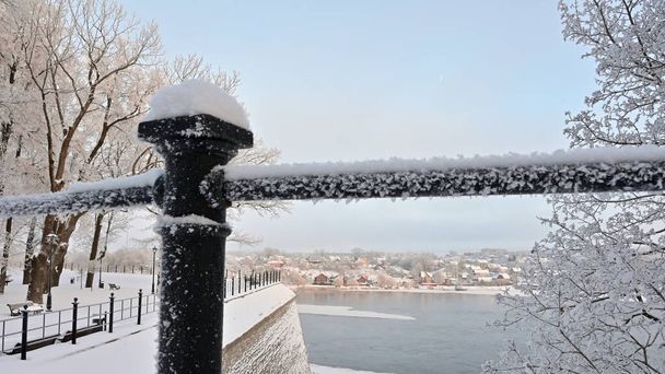 Through a metal fence richly covered with hoarfrost, a beautiful view of the snow-covered park on top of the historical bastion, the unfrozen river and the one-story city under a blue sky with a light haze opens up. - Photo, Image