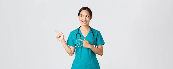Covid-19, gezondheidswerkers, pandemisch concept. Intrigued mooie aziatische vrouwelijke stagiaire, verpleegster in scrubs kijken en wijzen linksboven hoek met nieuwsgierige dromerige glimlach, witte achtergrond - Foto, afbeelding