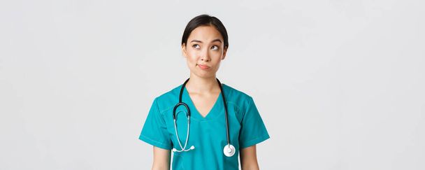 Egészségügyi dolgozók, vírusmegelőzés, karantén kampány koncepció. Átgondolt ázsiai női orvos, ápolónő köpenyben néz a bal felső sarokban, és vigyorog bizonytalan, gondolkodás, fehér háttér - Fotó, kép