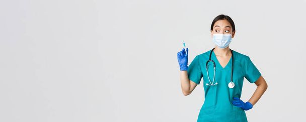 Covid-19, maladie du coronavirus, concept des travailleurs de la santé. Infirmière asiatique excitée avec masque médical, gants et gommages en caoutchouc, regardant avec étonnement la seringue avec vaccin, fond blanc - Photo, image